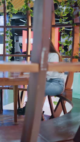 dama sentada con las piernas cruzadas en una mesa de madera en una elegante zona de bar interior, rodeada de vegetación e iluminada por luces decorativas, creando una atmósfera de moda y relajante