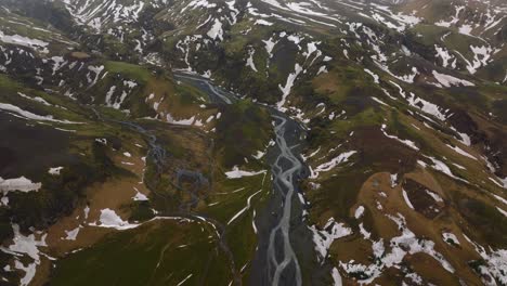 Aerial-panoramic-view-of-a-river-flowing-through-mountains-covered-in-melting-snow,-on-a-cloudy-and-foggy-day,-in-Iceland