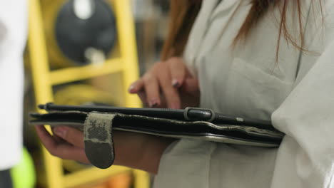 close-up of lady with polished nails using tablet in mechanical workshop, blurred background with industrial equipment and workspace, suggesting technical documentation and digital research