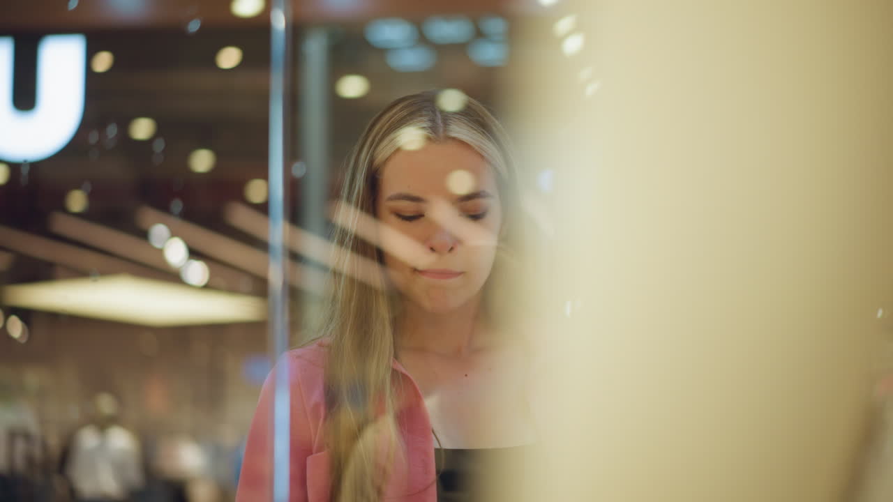 Lady in pink cloth gazes through glass with a contemplative expression, reflecting blurred lights and a glowing white sign in the background, the glass reflects soft glowing lights