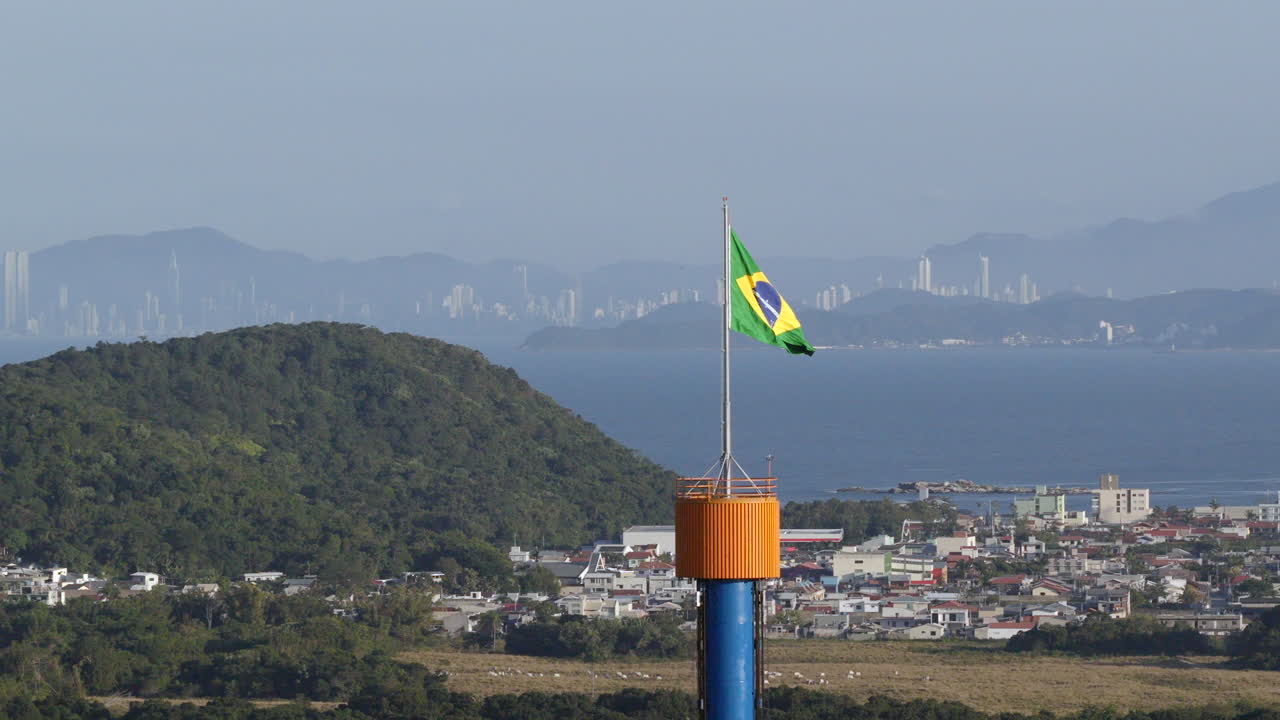 Brazilian flag waving in Penha, Santa Catarina, with Balneário Camboriú’s skyline visible across the bay, highlighting the Atlantic coast and surrounding cityscape, Slow-motion aerial descending shot