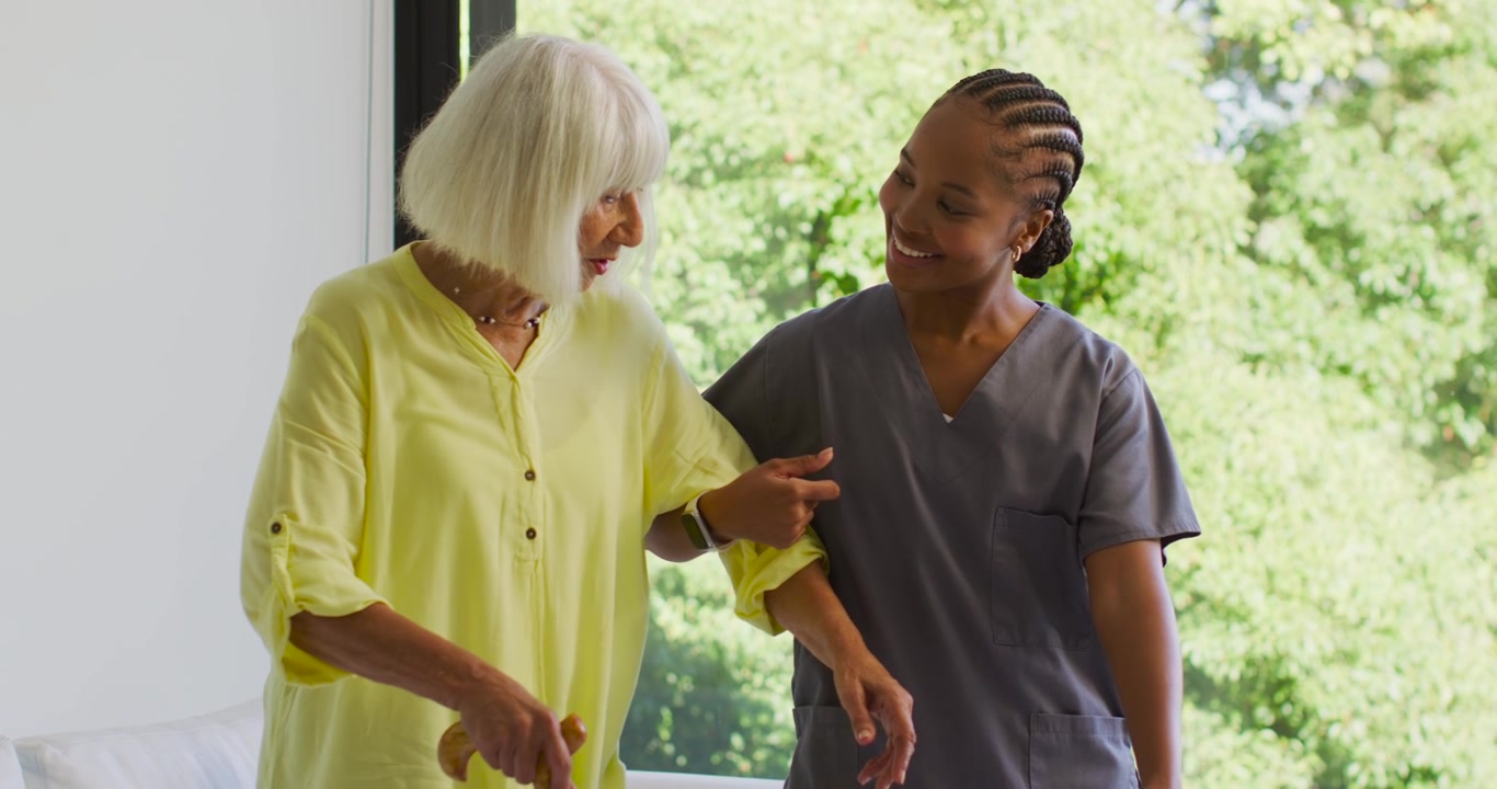 Senior with African American nurse guiding senior using cane near window to assist walking