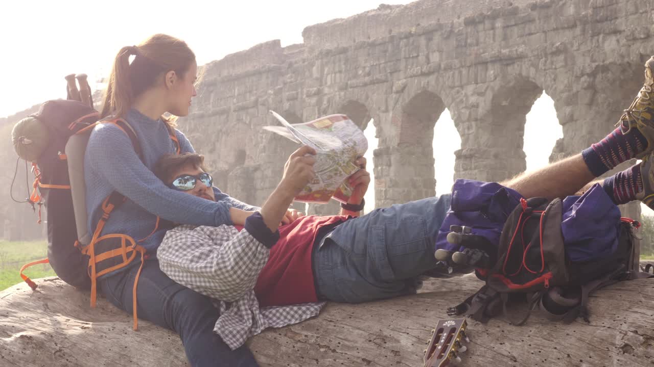 pareja feliz de mochileros turistas sentados acostados en un tronco de madera leyendo una guía de mapas apuntando a las antiguas ruinas del acueducto romano en el parco degli aquedotti parque en roma al amanecer romántico con saco de dormir cámara lenta