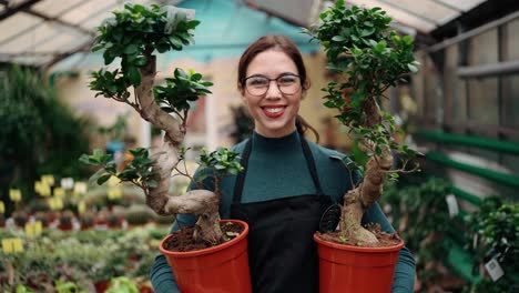 Young-female-florist-in-apron-walking-among-rows-of-flowers-in-flower-shot-or-greenhouse-while-holding-two-pots-with-plants