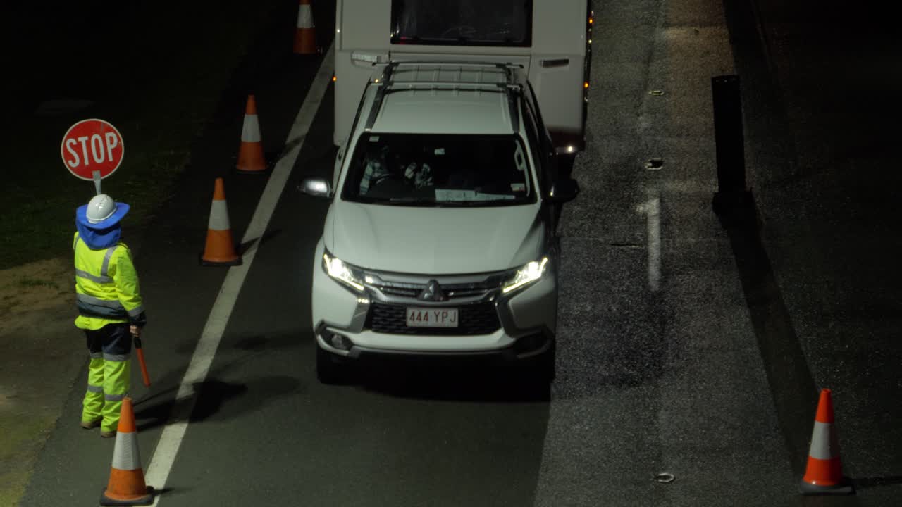 Traffic Guard In Reflective Uniform Giving A Go Signal On Cars Travelling At Night - Traffic At The NSW - QLD State Border Checkpoint - Gold Coast, Australia. - high angle shot
