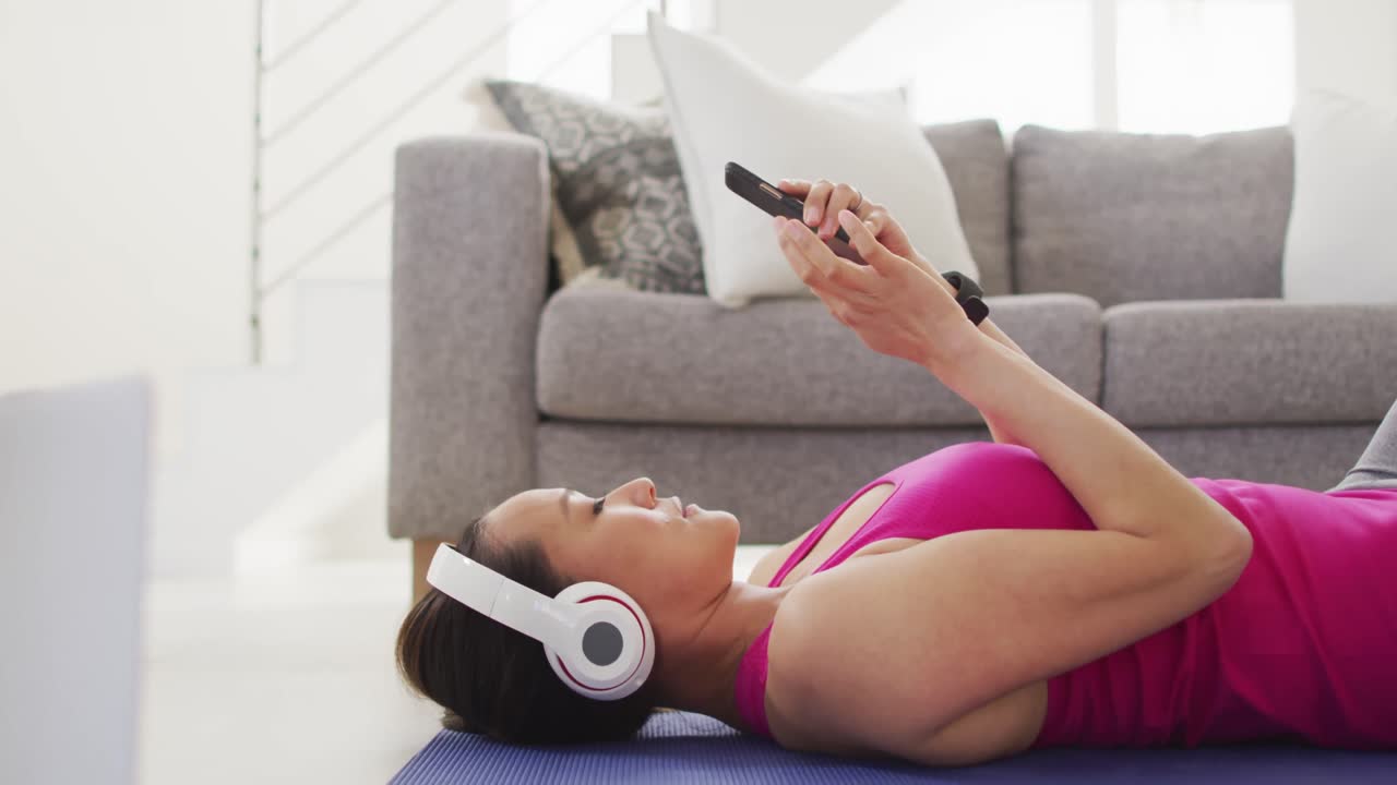 mujer asiática con ropa de fitness acostada en un tapete con auriculares, usando un teléfono inteligente en casa