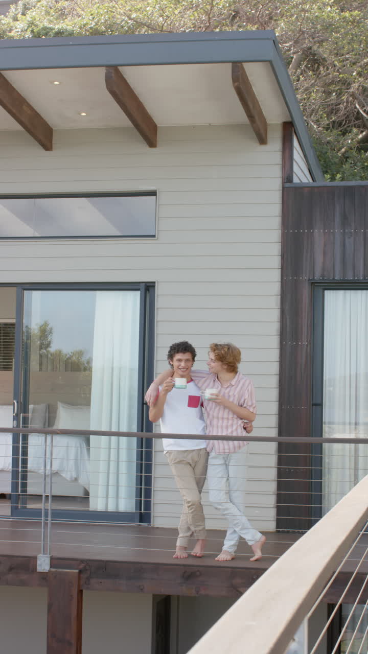 Vertical video of happy diverse gay male couple drinking coffee on balcony at home, slow motion