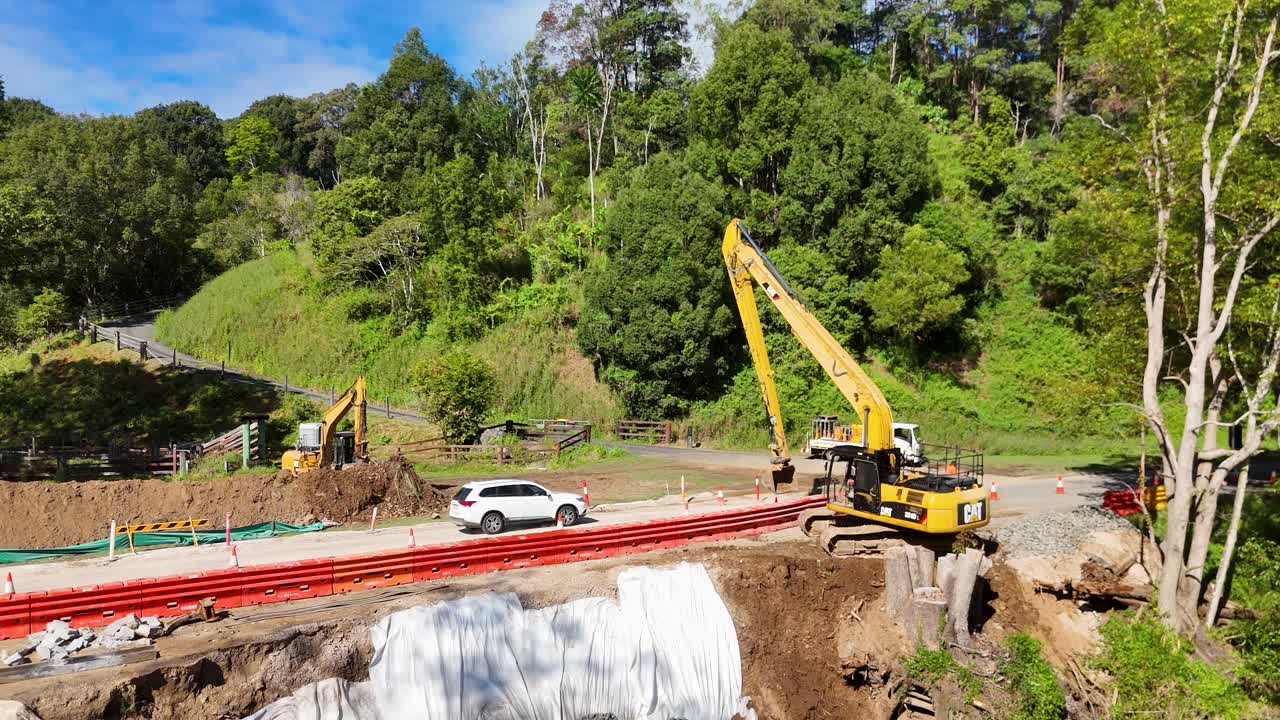 Aerial view of roadwork with an excavator near a river in Uki, NSW, Australia. Bright daylight highlights the construction scene