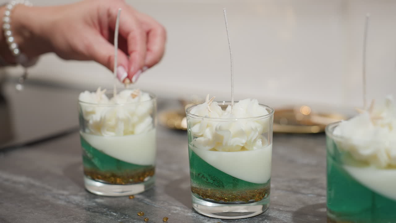 House wife carefully picks shell-like decorative element from table and gently places it on whipped top of candle jar with green and white layers, creating elegant handcrafted decorative centerpiece