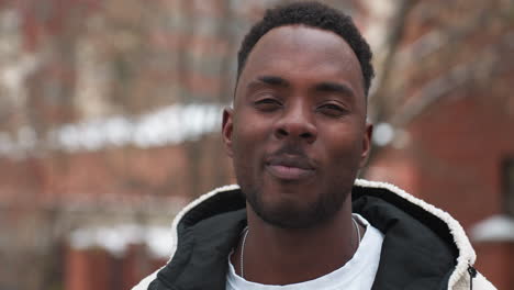 black celebrity with afro hairstyle wearing winter hoodie, giving warm confident smile outdoors with trees and buildings in background, radiating positive energy and style