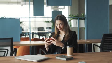 una mujer joven y bonita con ropa de negocios se sienta en una elegante oficina moderna de coworking, jugando a un juego en su teléfono inteligente alegremente y atentamente