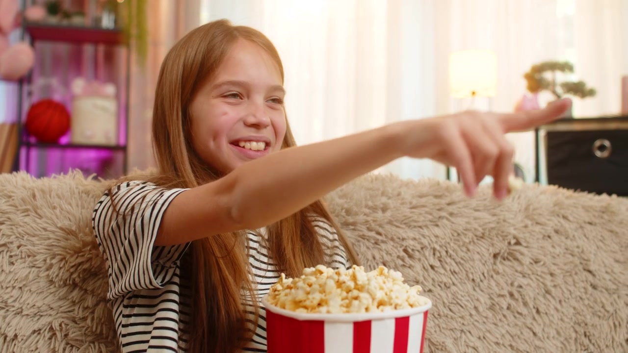 Barn pige spiser glad popcorn mens hun ser tegnefilm på tv og griner muntert i sofaen