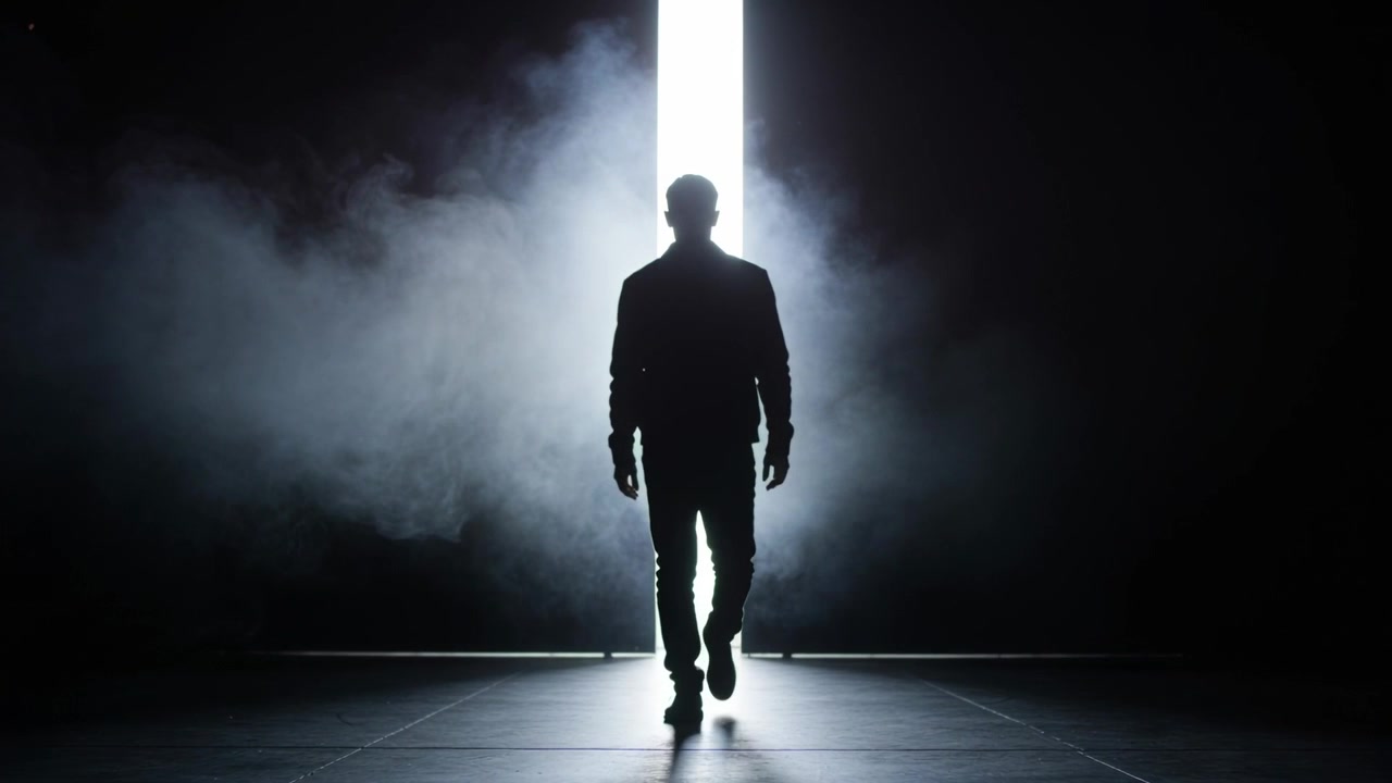Uomo che emerge camminando da una fessura di luce su un pavimento piastrellato attraverso la nebbia verso la telecamera, in giacca scura.