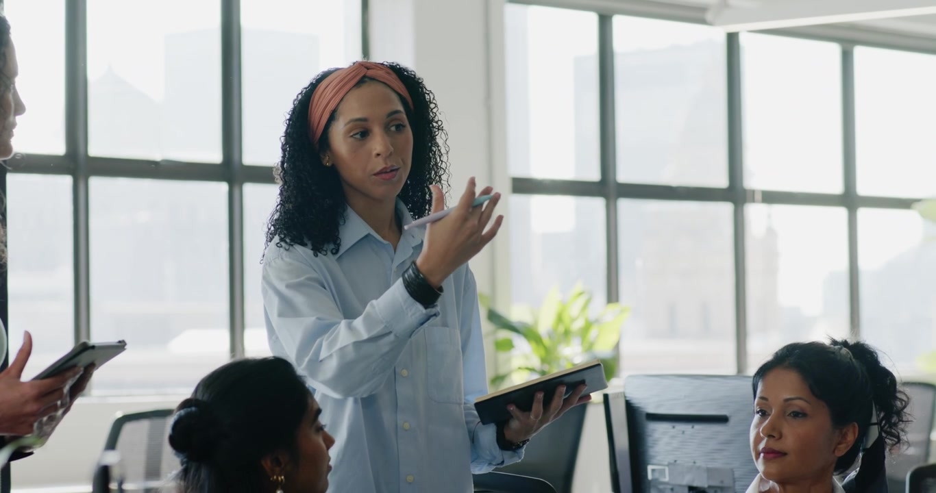 Diverse female colleagues watching team lead rising from seat presenting plan at office with tablet