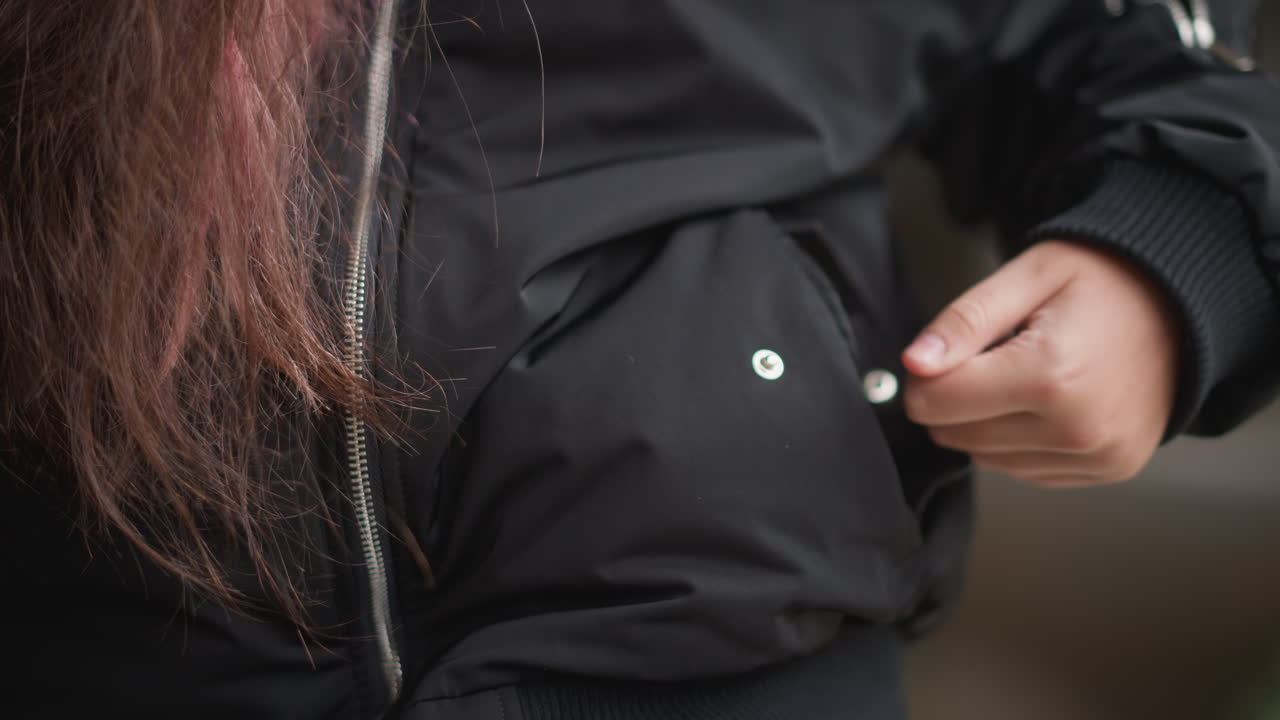 Wet black jacket hand in pocket, young woman commuter standing in drizzle with droplets on sleeve and pink hair, closeup of zipper and pocket, reflective puddle nearby, moody urban atmosphere