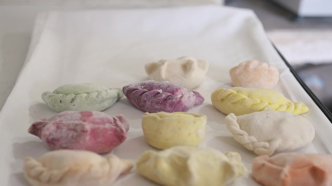 Traditional Argentine empanadas with multicolored dough varieties displayed frozen on preparation tray, featuring handmade folded pastry textures and assorted savory fillings, slow reveal arc shot.