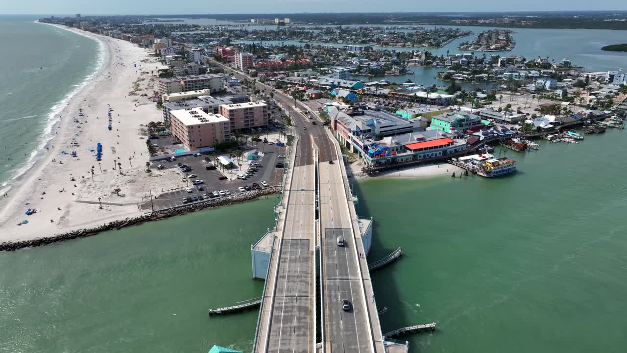 Aerial backwards shot of driving cars on John's Pass Bridge in Florida at sunny day. Wide shot. Sandy beach with relaxing people in Madeiras beach City. Coastline luxury houses and islands in back.