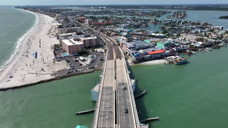 aerial backwards shot of driving cars on john's pass bridge in florida at sunny day. wide shot. sandy beach with relaxing people in madeiras beach city. coastline luxury houses and islands in back.