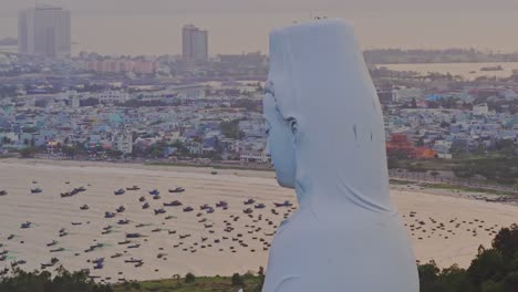 Majestic-Lady-Buddha-Statue-from-behind,-overlooking-the-city-of-Da-Nang-and-the-ocean-filled-with-numerous-boats