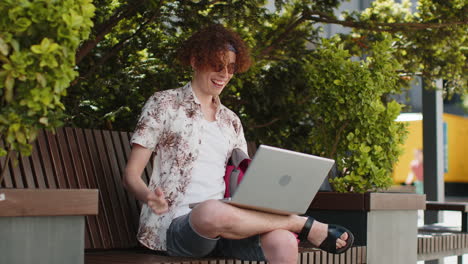 Happy man tourist using laptop computer celebrating win good message news sitting on bench in city