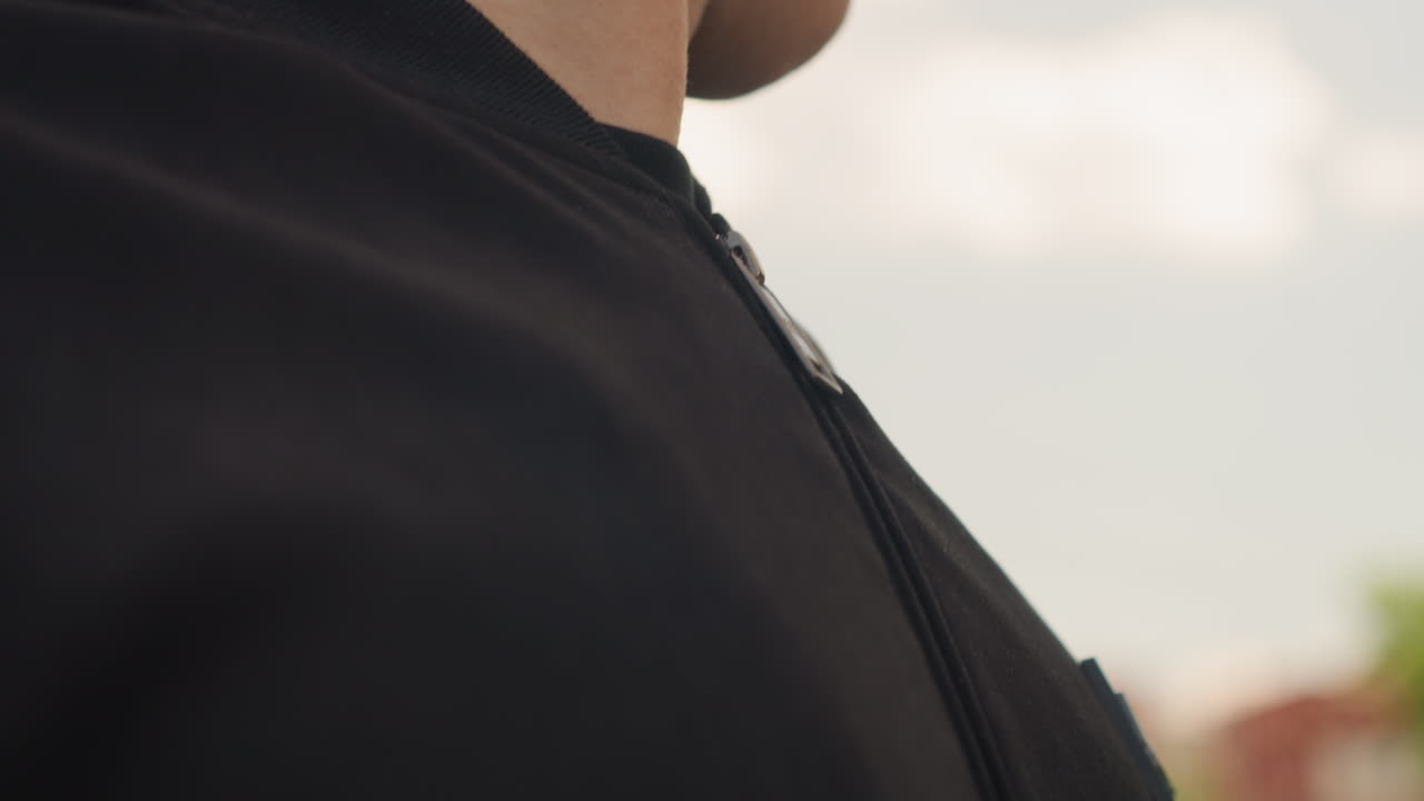 남자가 야외에서 목 부분의 코트를 여미고, 남자가 야외 환경에서 옷깃 지퍼를 조절하며, 남자가 바람이 부는 날씨에 야외에서 옷깃 근처의 지퍼를 잠그며 재킷을 준비하고 있어요.