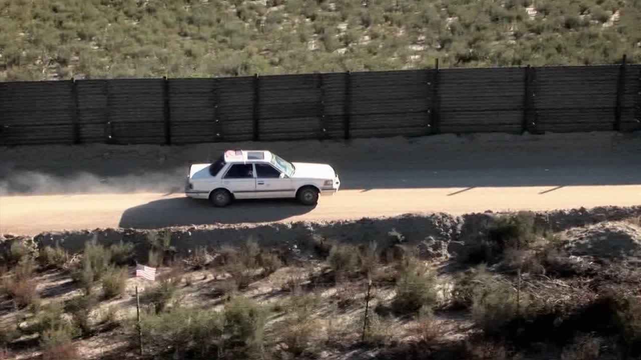 선루프가 달린 흰색 차가 높은 나무 울타리로 둘러싸인 사막 도로를 따라 운전하고 있습니다.