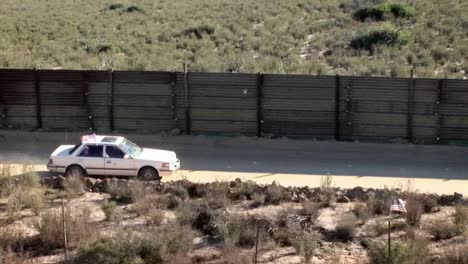 a white car with a sunroof is driving along a desert road bordered by a tall wooden fence