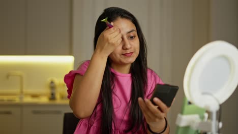 una chica morena feliz se aplica polvo a la cara y se maquilla en un vestido rosa durante su video de blog de belleza sobre apariencia, belleza y cosméticos en un apartamento moderno durante el día