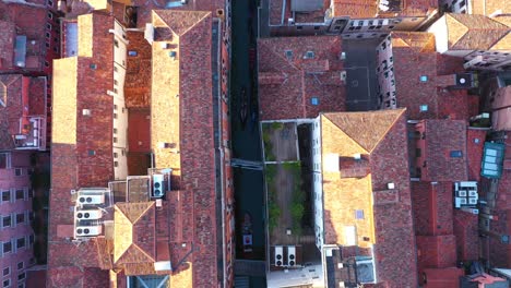 Vista-Aérea-De-Arriba-Hacia-Abajo-Volando-Sobre-El-Canal-Y-Los-Techos-Mientras-La-Góndola-Pasa-En-Un-Día-Soleado-En-Venecia-En-Italia-En-4k