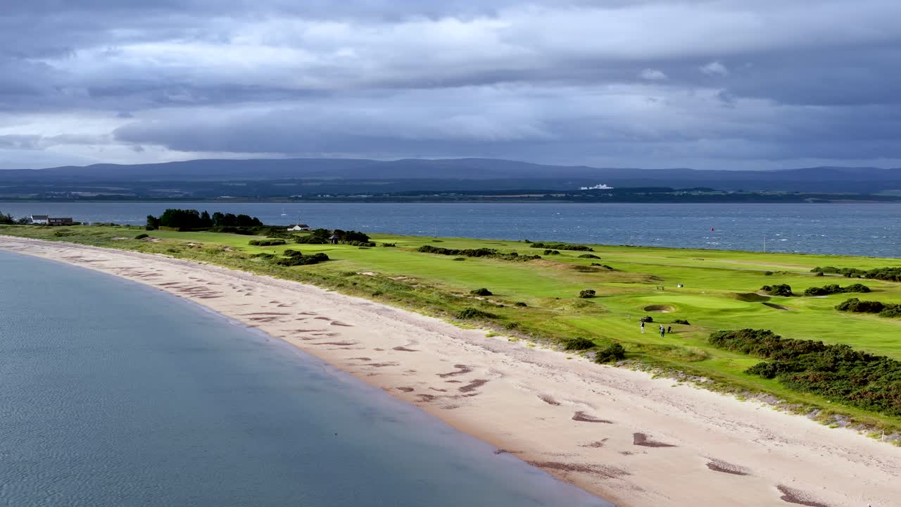 Drone glides above sandy beach, green fields, and calm sea under dramatic, cloudy summer sky