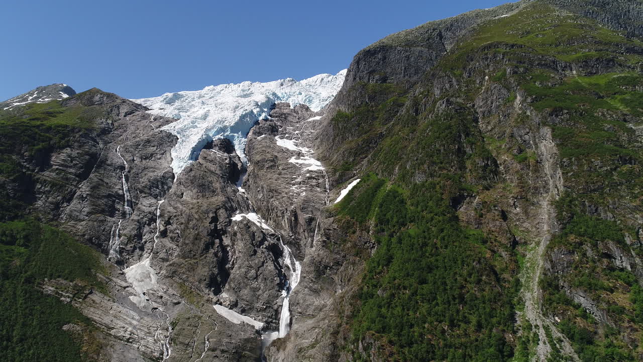드론은 은 파란 하늘 아래 빙하 폭포와 푸른 식물이 있는 바위 절벽에 있는 빙하를 촬영합니다.