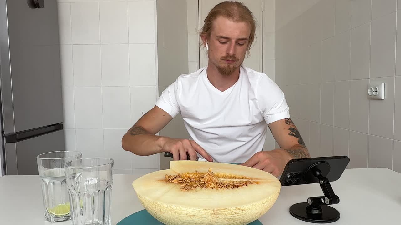 Mand spiser frisk melon og ser på telefon i køkkenet.