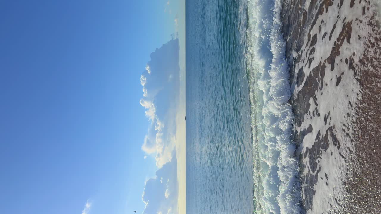 Immagine verticale di una spiaggia tranquilla senza persone, schiuma bianca, piccole pietre, spazio per il testo su cielo blu.