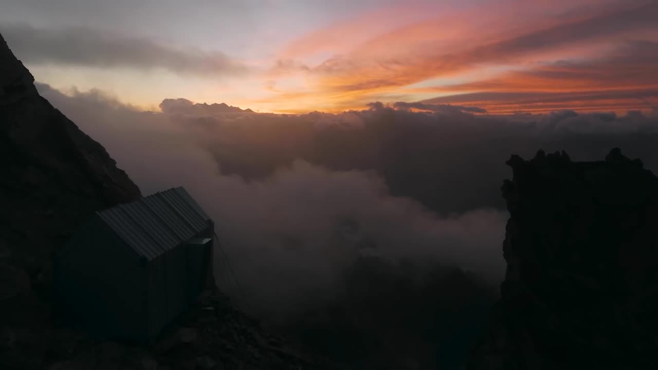 Aerial drone flying over clouds and mountains in reverse at Western Alps in Valle D'Asota during sunset or sunrise with sky being orange on top of clouds. A small cabin for climbers is on the cliff.