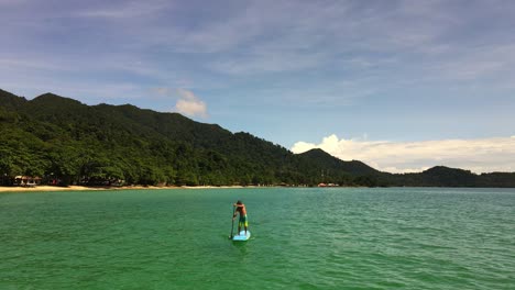 low angle aerial drone bird's eye view of caucasian man exercising on a sup paddle board in turquoise tropical clear waters, with beach and coastline in thailand