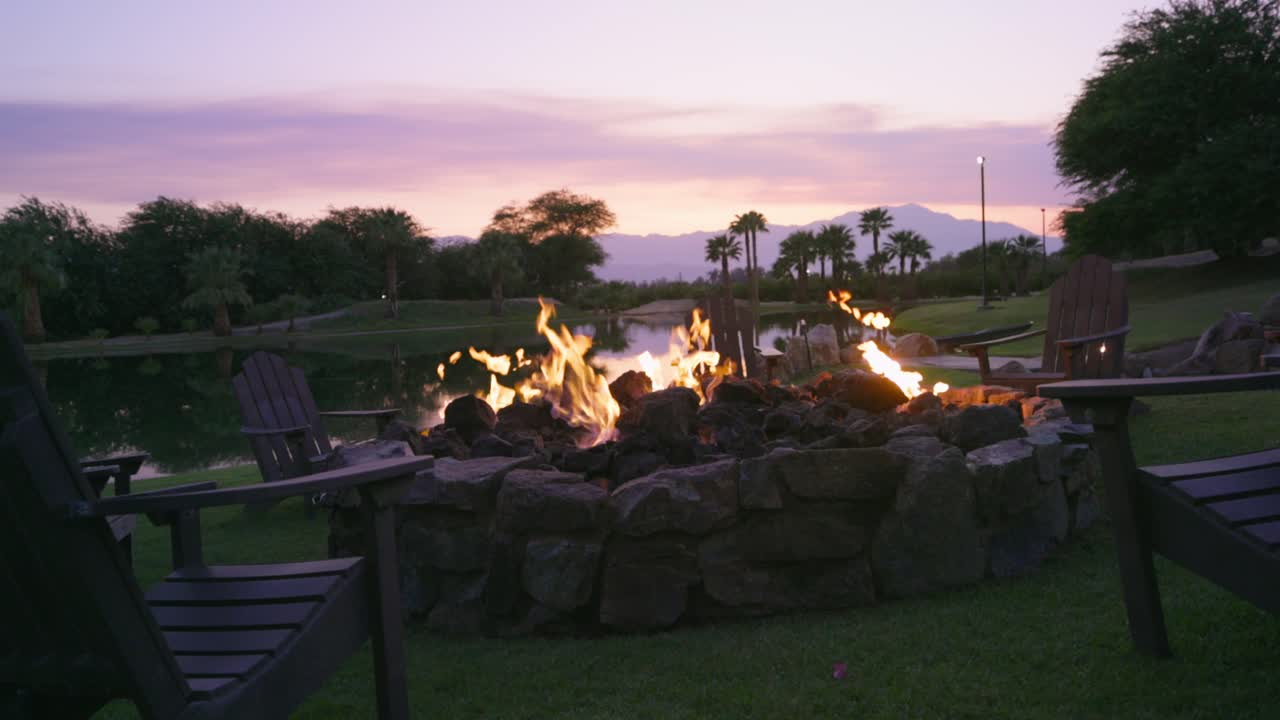 Several relaxing chairs around a large modern gas powered bonfire on a lake, while the sky in the background is beautifully colored by the setting sun behind the palm trees. slow mo shot