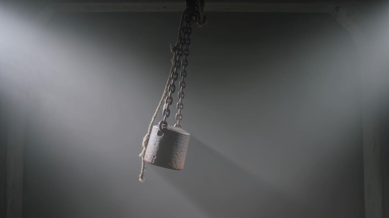 Swinging metal cylinder rotating from initial tilt in dim warehouse, with chains and frayed rope