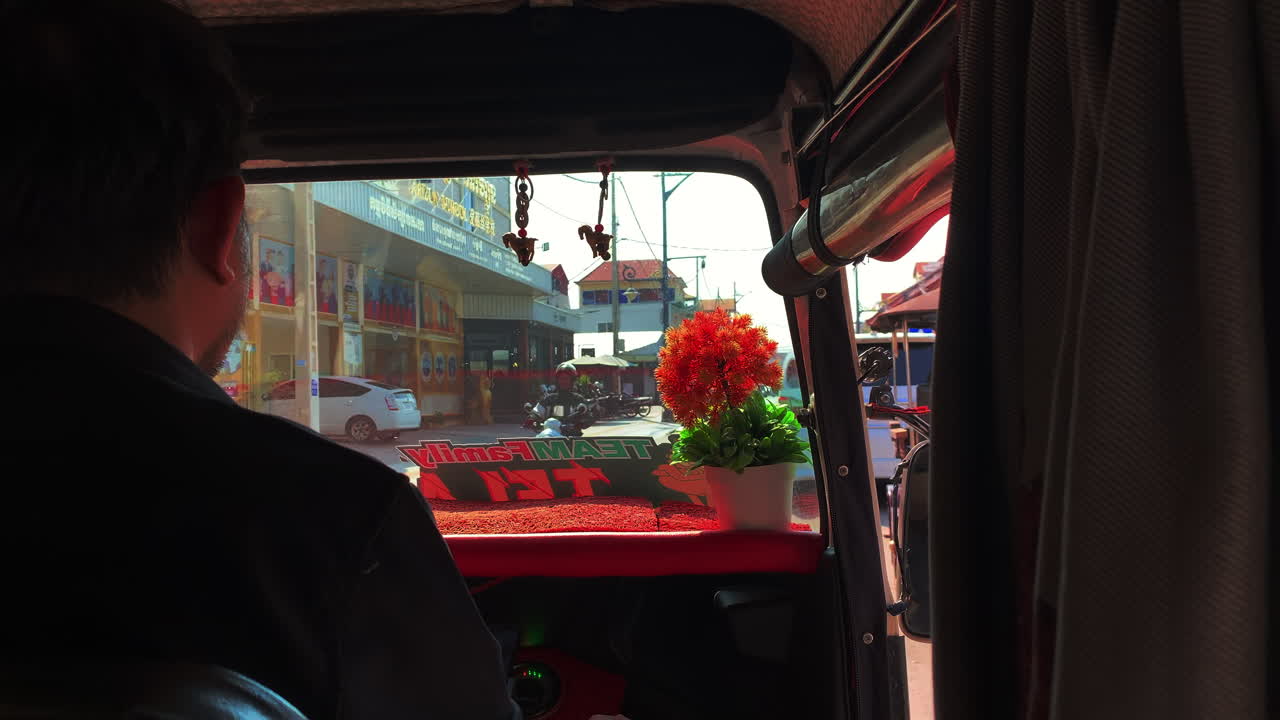 Tuk-Tuk driver with flowerpot on dashboard and hanging pendants turns left past buildings and shops on a city street, moving shot