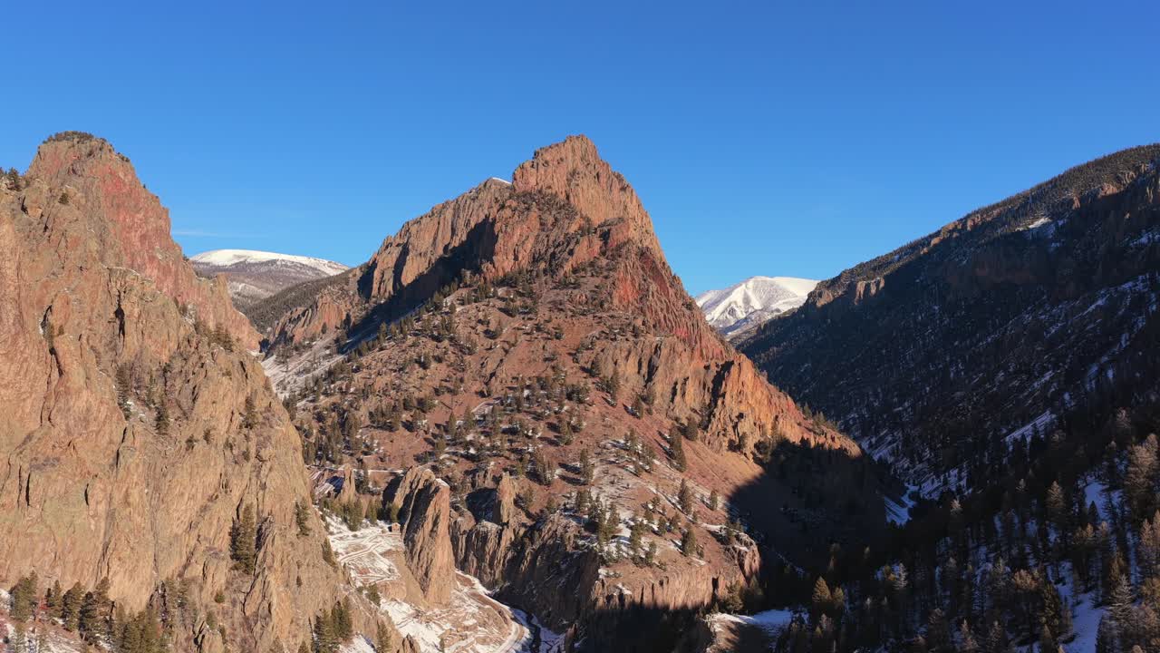 Una vista aerea cinematografica mette in risalto un'imponente cima montuosa che si erge sopra ripide pareti rocciose del canyon. Ombre profonde e drammatiche e neve invernale mostrano l'immensa scala di questo aspro ambiente montano.