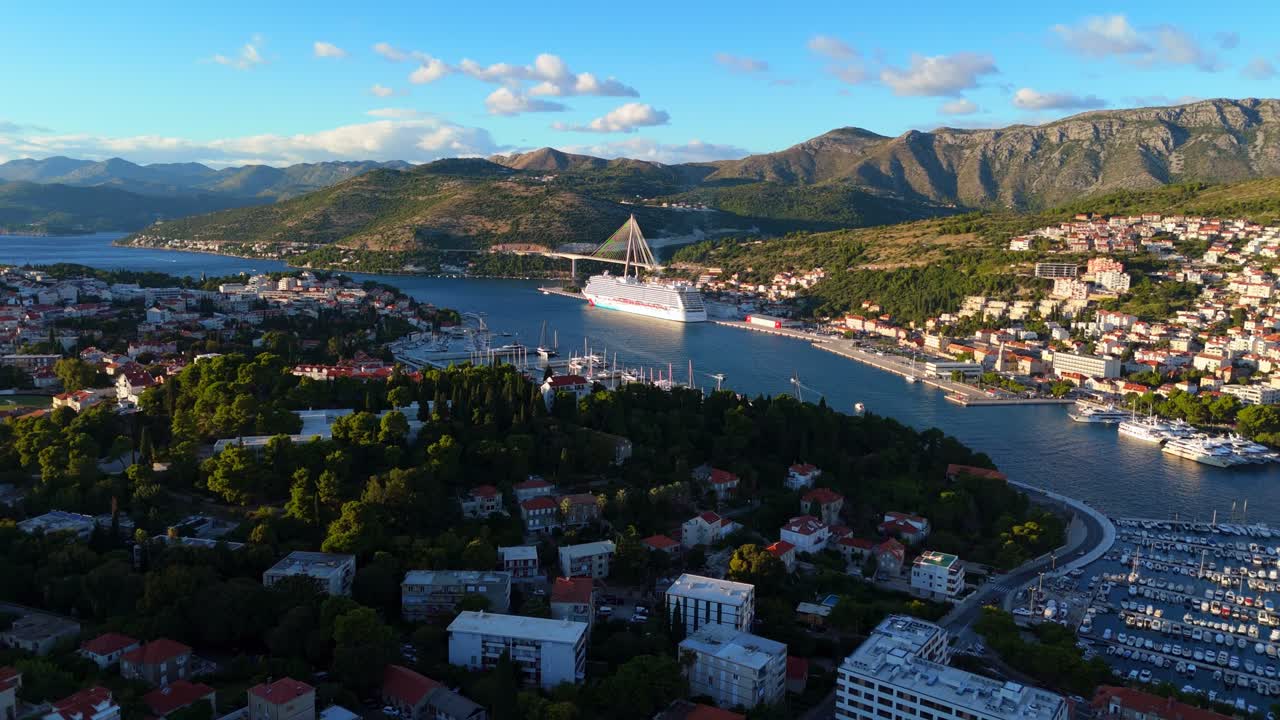 Aerial wide-angle drone captures Dubrovnik city and its cruise ship port, with majestic mountains rising in the background, as the golden sunset casts warm light over terracotta rooftops and harbour