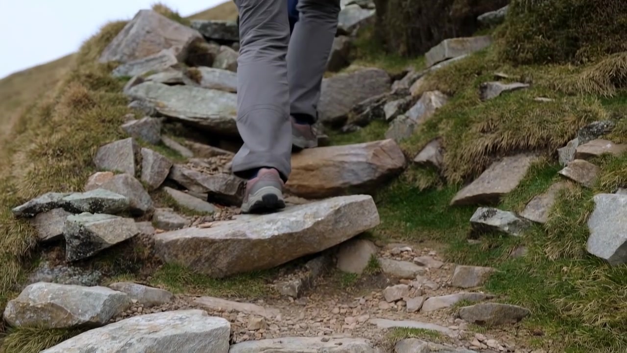 Scarponi da trekking che salgono un sentiero di montagna roccioso, primo piano delle gambe inferiori che scavalcano gradini di pietra, bordi muschiosi