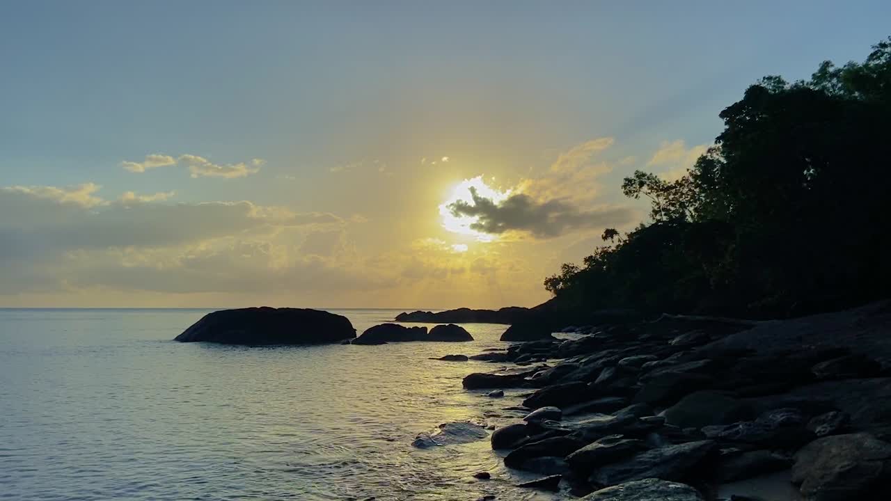 View along a rocky shoreline with silhouetted headland and rocks, as the dawn sun breaks through the clouds on the horizon in a blue and gold sky, as the gentle waves lap at the shore