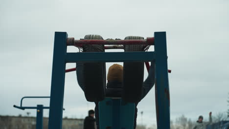 back view of a coach exercising on outdoor equipment, lifting weights with red bars as stops and moves his body and lifts his right leg, while two boys are working out on a track