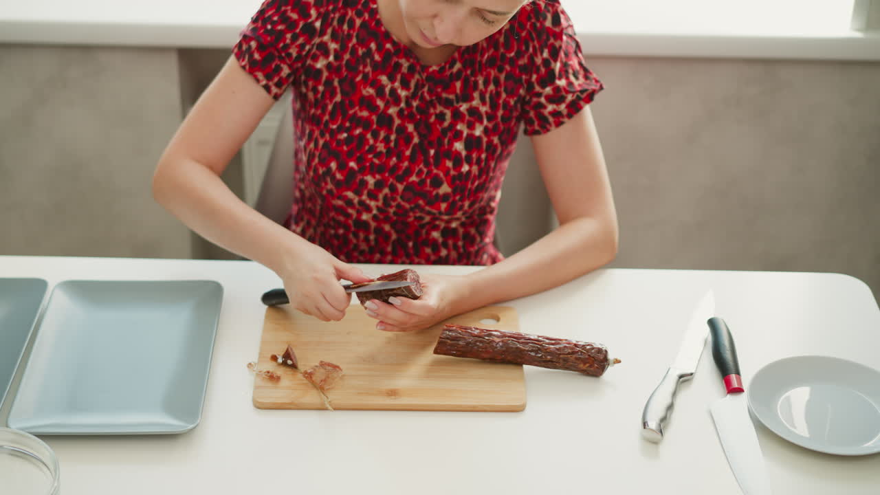 Young woman seated by window, using knife to peel casing from dried sausage with focused effort, wooden board and minimal table setup under soft daylight highlighting cooking prep technique
