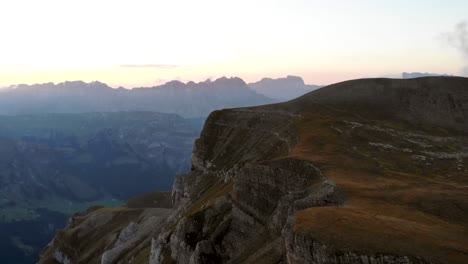 Aerial-view-of-the-cliffs-and-mountains-of-Linthal-in-Glarus,-Switzerland-during-sunset-with-pan-down-towards-the-valley