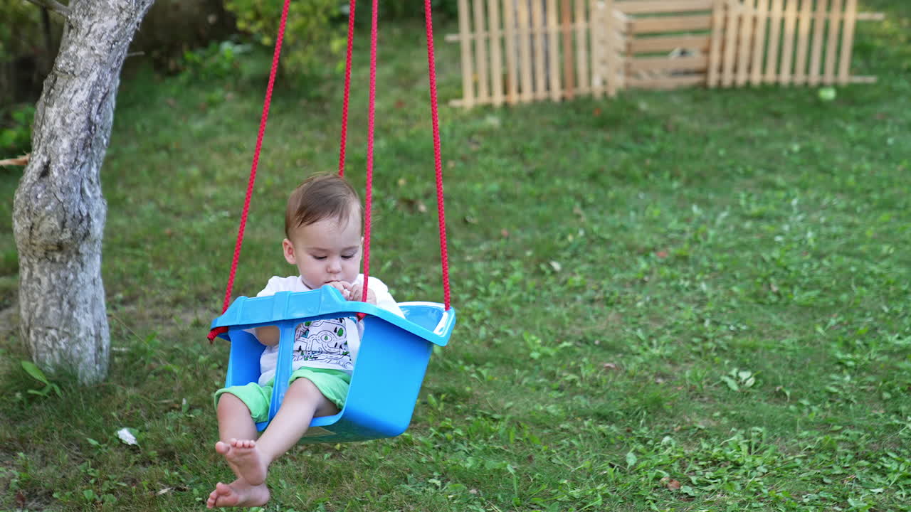 Little boy swaying in a swing looking attentively at the fastener in his hands. Cute kid in the summer garden. Nature backdrop.