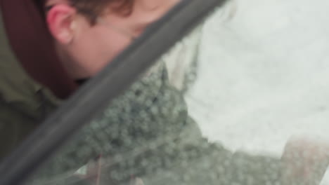 man walks out from red car and closes door as snowfall settles on vehicle window, captured through glass with water droplets distorting view, highlighting cold weather