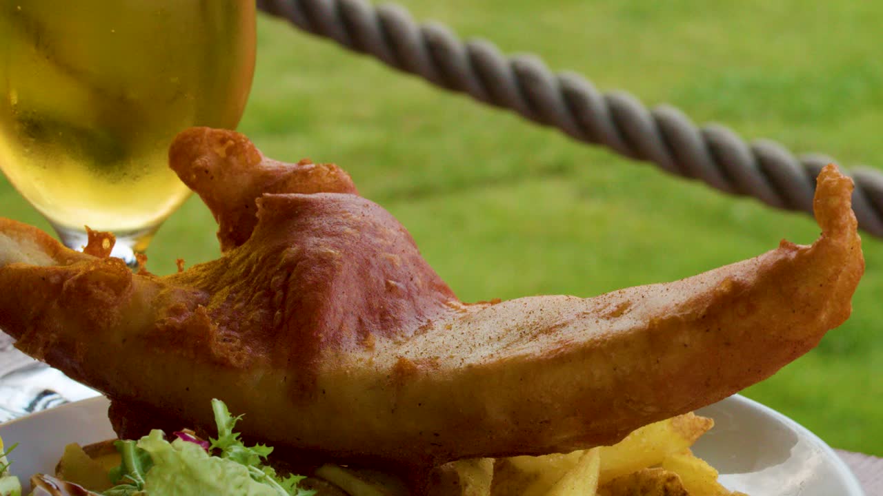 Hand squeezes lemon onto crispy fish and chips beside beer, shot outdoors in natural daylight
