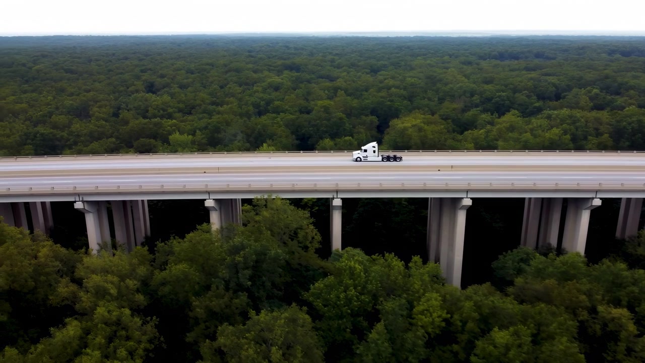 Side tracking view follows white highway tractor moving across long concrete viaduct, while layered treetops fill surrounding landscape beneath soft daylight and pale sky.