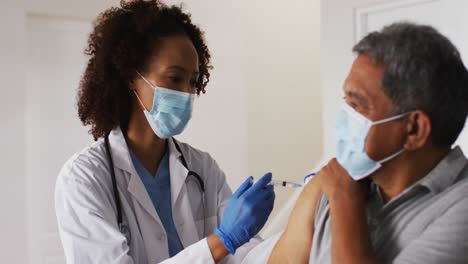 Mixed-race-female-doctor-wearing-mask-giving-vaccination-to-senior-man-at-home